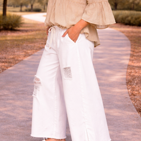 side view of woman wearing distressed white wide leg jeans with flowy off the shoulder boho blouse and flat sandals walking outdoors
