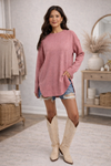 Woman wearing an oversized dusty rose cozy cardigan, denim shorts and cream tall boots in a cozy room.