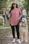 A woman wearing a dusty rose versized fall sweater, black leggings, sneakers, and a ball cap walking a dog in park setting with other people and dogs.