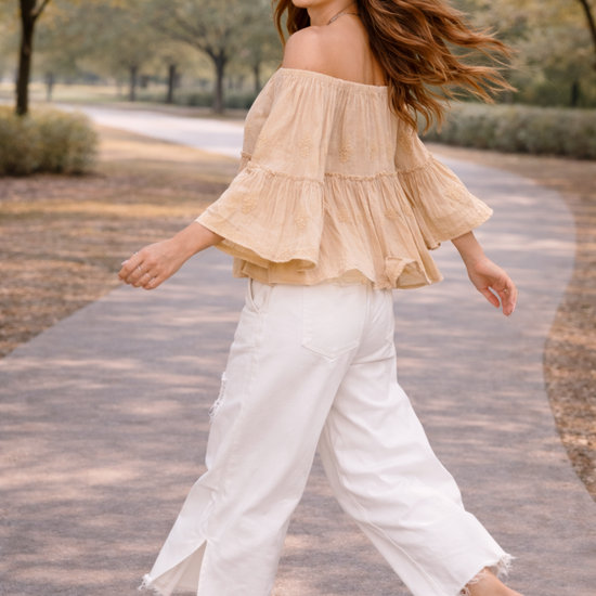 Woman walking with movement in a neutral boho off-shoulder blouse and distressed white wide-leg jeans with sandals, styled in a soft golden-hour outdoor setting