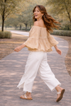 Woman walking with movement in a neutral boho off-shoulder blouse and distressed white wide-leg jeans with sandals, styled in a soft golden-hour outdoor setting