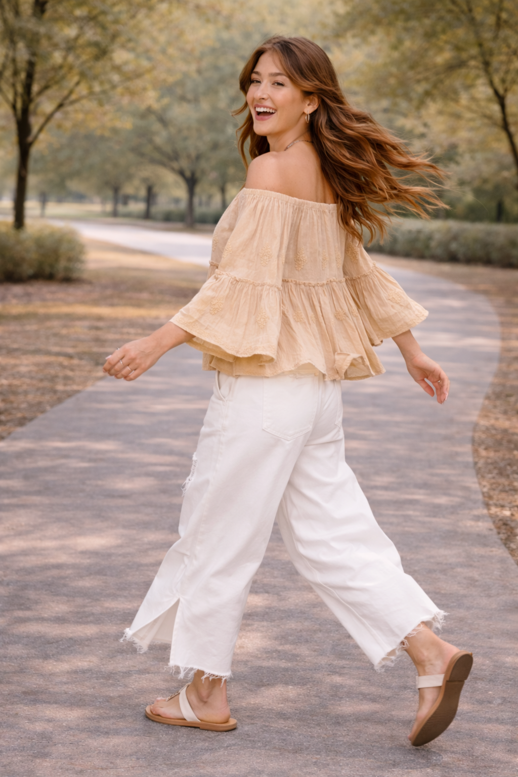 Woman walking with movement in a neutral boho off-shoulder blouse and distressed white wide-leg jeans with sandals, styled in a soft golden-hour outdoor setting