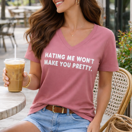 Woman sitting at an outdoor café wearing a dusty rose Bella Canvas V-neck graphic tee with the phrase “Hating me won’t make you pretty,” styled with denim shorts and holding an iced coffee