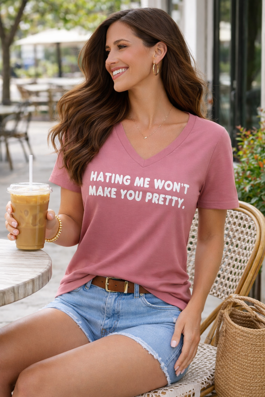 Woman sitting at an outdoor café wearing a dusty rose Bella Canvas V-neck graphic tee with the phrase “Hating me won’t make you pretty,” styled with denim shorts and holding an iced coffee
