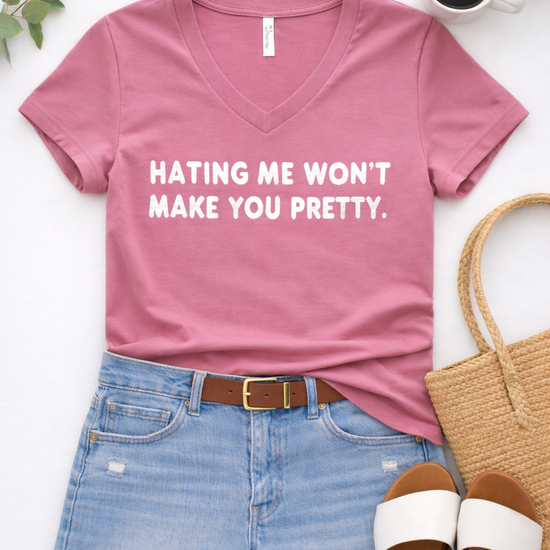 Flat lay of a dusty rose Bella Canvas V-neck graphic tee with “Hating me won’t make you pretty” paired with distressed denim shorts, a brown belt, white slide sandals, and a woven tote