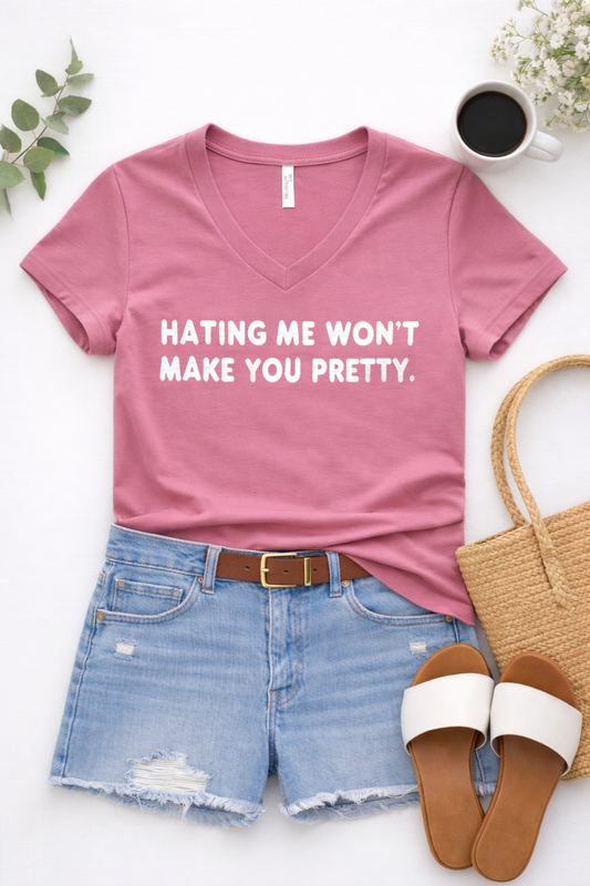 Flat lay of a dusty rose Bella Canvas V-neck graphic tee with “Hating me won’t make you pretty” paired with distressed denim shorts, a brown belt, white slide sandals, and a woven tote