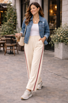 Woman styling ivory wide leg athleisure pants with crimson stripe with white tank top, denim jacket, and sneakers in casual everyday outfit
