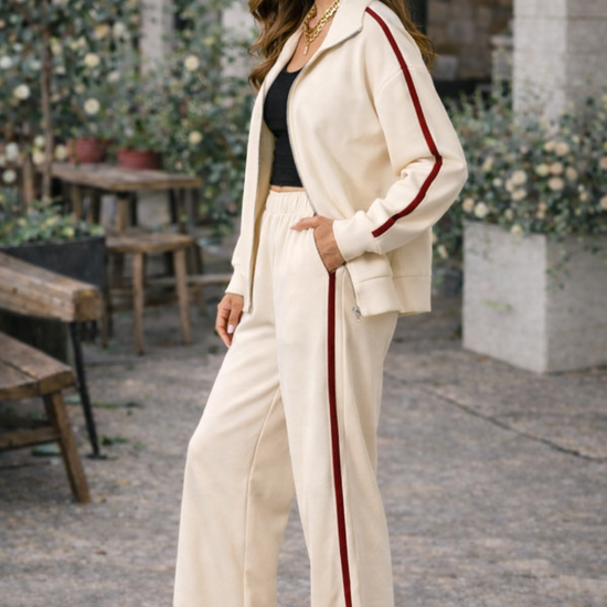 Side view of ivory wide leg athleisure pants with crimson stripe styled with matching track jacket, black crop top, and sneakers in casual café lifestyle setting