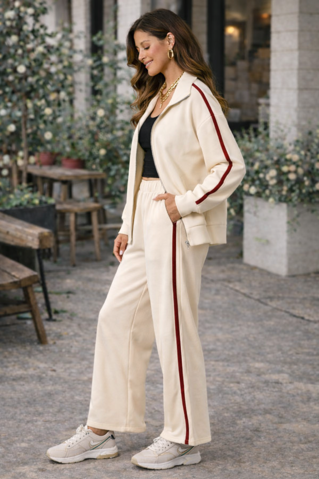 Side view of ivory wide leg athleisure pants with crimson stripe styled with matching track jacket, black crop top, and sneakers in casual café lifestyle setting