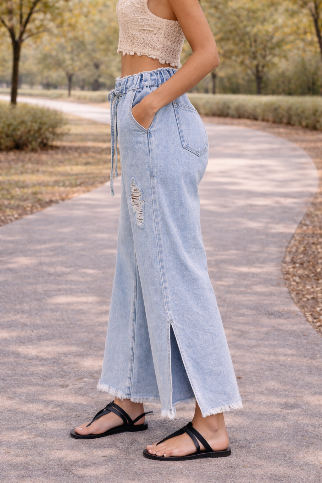 Side view of light wash distressed wide leg jeans with paper bag waist and drawstring detail, styled in a casual outdoor setting