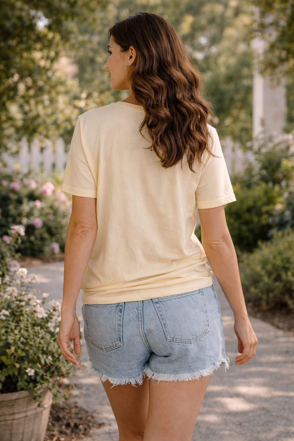 back view of woman wearing mama goose graphic tee with denim shorts walking on a sunlit garden path