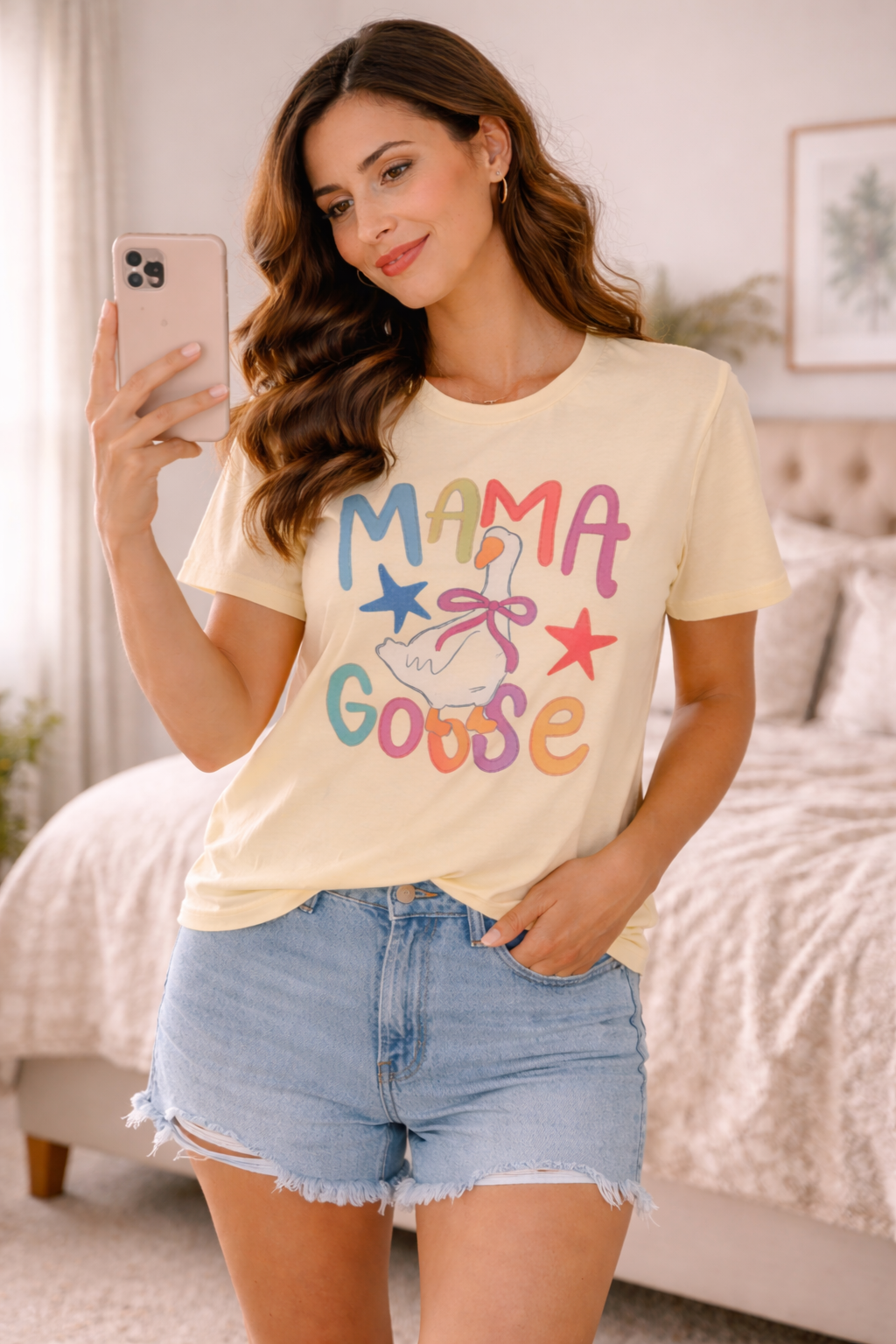 woman wearing mama goose graphic tee taking mirror selfie in a cozy bedroom wearing denim shorts