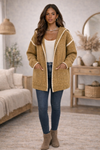 Woman wearing a mustard hooded boho cardigan with trim, jeans, ankle boots and a white tank top in a cozy living room.