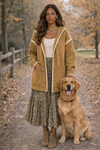 Woman wearing a white top, flowery skirt and a mustard hooded boho cardigan, in an outside fall setting with a golden retriever