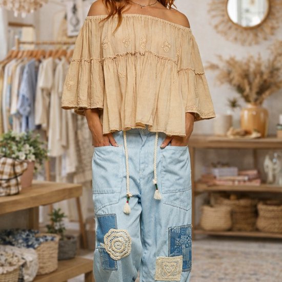 Woman wearing an off shoulder peasant top with sky blue boho patchwork wide leg pants in a store setting.