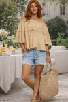 Woman wearing an off shoulder boho peasant flowy top with distressed denim, carrying an oversized straw bag in an outside setting.