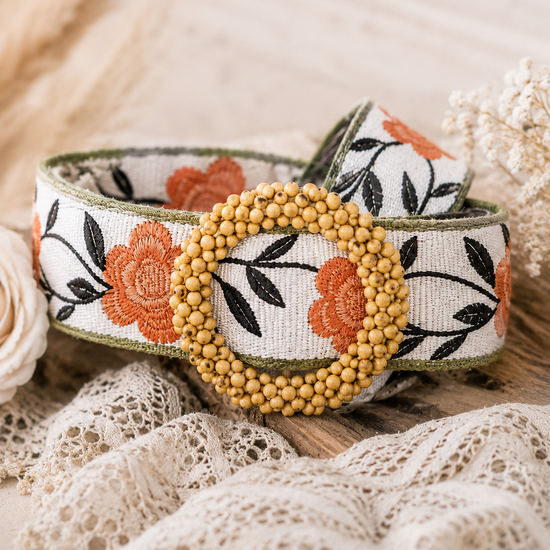 Close-up of the POL Boho Floral Statement Belt featuring orange floral embroidery, black leaf detailing, and a round wooden beaded buckle on a soft neutral boho background.