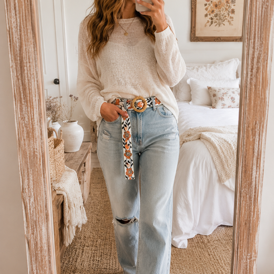Woman styling the POL Boho Floral Statement Belt with Beaded Buckle in a cozy mirror selfie wearing a cream sweater and light wash denim jeans.