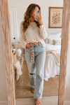 Woman styling the POL Boho Floral Statement Belt with Beaded Buckle in a cozy mirror selfie wearing a cream sweater and light wash denim jeans.