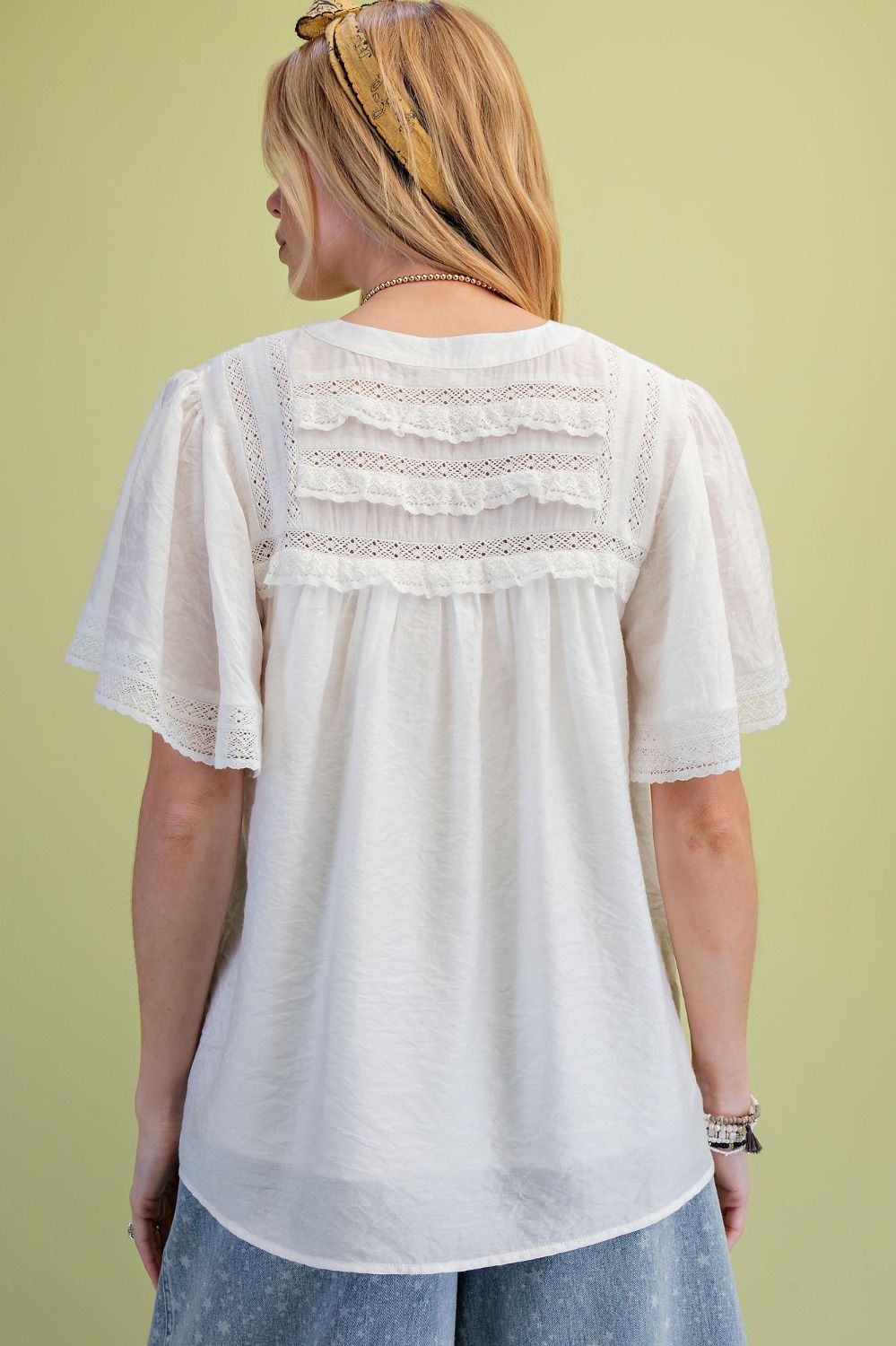 A model is standing in front of a green background showing the back view of the Ivory Lace Pintuck Flutter Sleeve Blouse