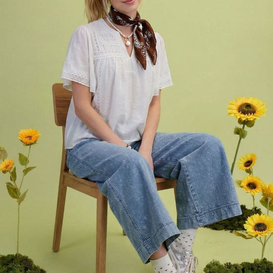 Lifestyle image of the Ivory Lace Pintuck Flutter Sleeve Blouse. The model is sitting on a stool with a green background