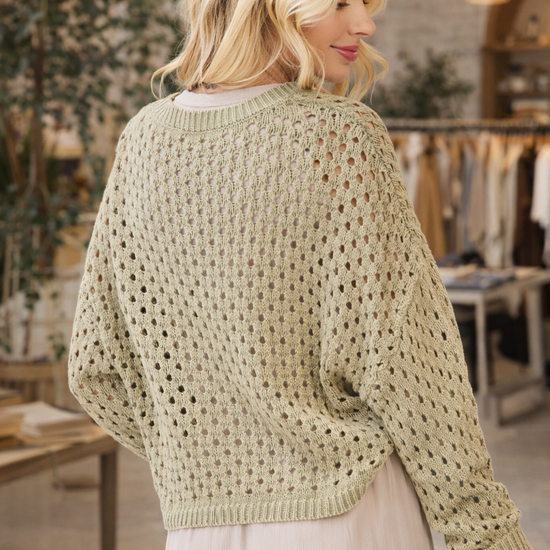 Back view of a woman wearing a sage green open knit long sleeve crochet sweater styled with a neutral skirt in a boutique setting – lightweight boho sweater for women