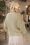 Back view of a woman wearing a sage green open knit long sleeve crochet sweater styled with a neutral skirt in a boutique setting – lightweight boho sweater for women