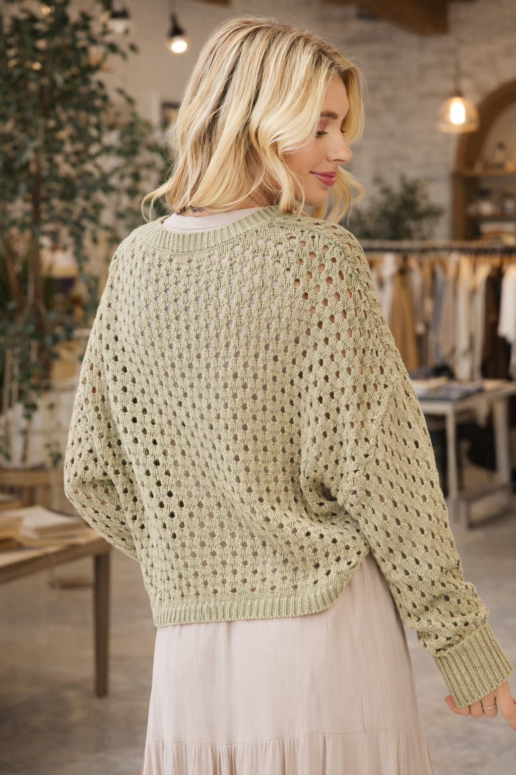 Back view of a woman wearing a sage green open knit long sleeve crochet sweater styled with a neutral skirt in a boutique setting – lightweight boho sweater for women