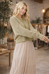 Woman wearing a sage green open knit long sleeve crochet sweater styled with a neutral tiered skirt in a boutique setting – lightweight boho sweater for women