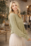 Woman wearing a sage green open knit long sleeve crochet sweater styled with a neutral skirt in a boutique setting – lightweight boho sweater for women