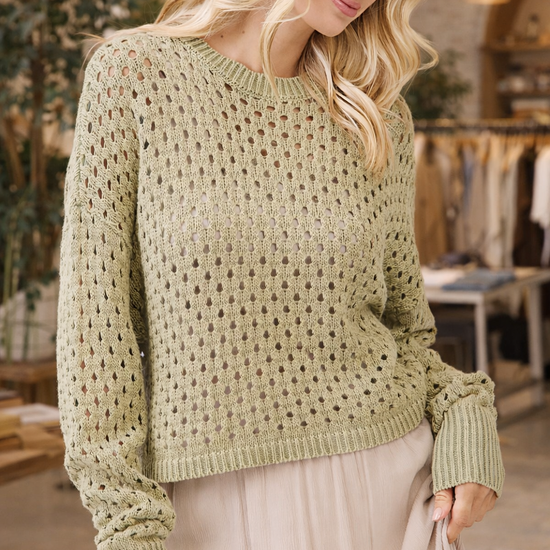 Woman wearing a sage green open knit crochet sweater styled with a neutral skirt in a boutique setting – lightweight boho sweater for women