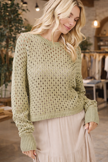 Woman wearing a sage green open knit crochet sweater styled with a neutral skirt in a boutique setting – lightweight boho sweater for women