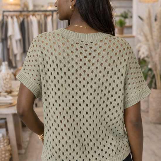 Back view of sage green open knit short sleeve sweater top on African American model inside boutique