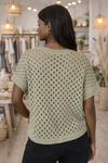 Back view of sage green open knit short sleeve sweater top on African American model inside boutique