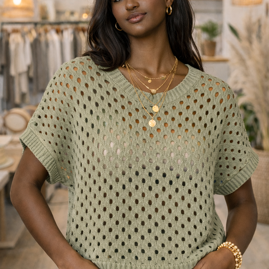 African American model wearing a sage green open knit short sleeve sweater top styled with black jeans in a neutral boutique setting.