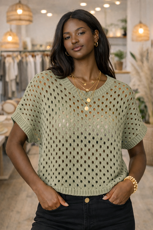 African American model wearing a sage green open knit short sleeve sweater top styled with black jeans in a neutral boutique setting.