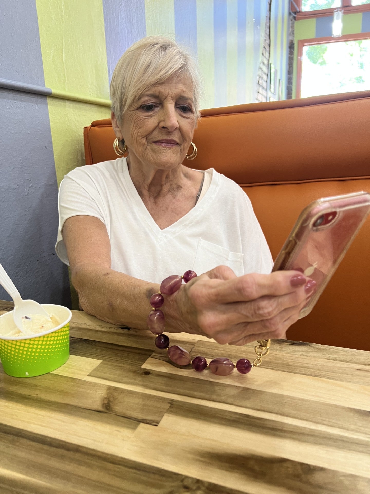 Woman wearing the shimmery berry orchid phone wristlet sitting at a table in an ice-cream shop