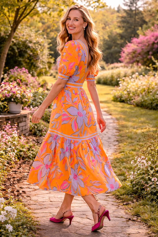 Side view of a woman walking along a sunlit garden path wearing an orange floral midi dress with puff sleeves and a flowy tiered skirt, styled for a bright spring and summer look