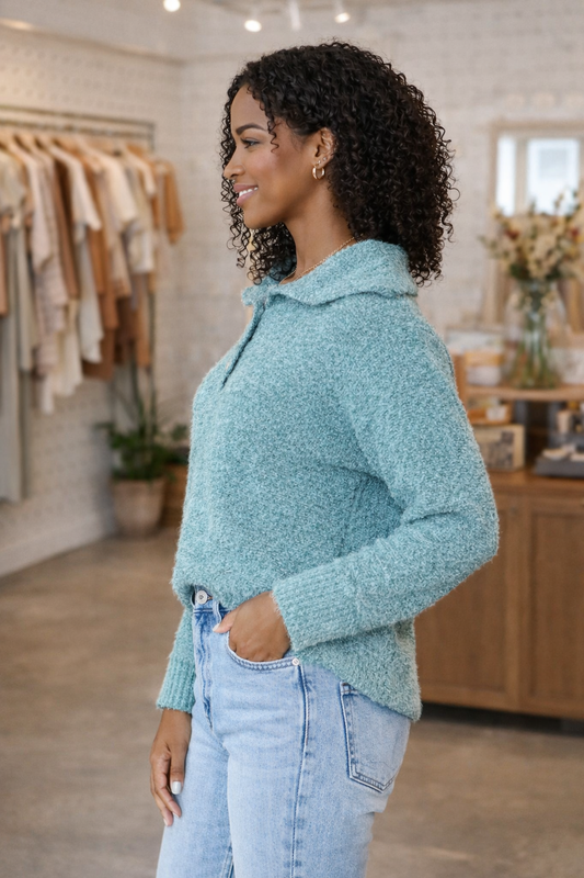 side view of African American woman wearing a teal textured boucle sweater styled with light wash jeans in a neutral boutique setting