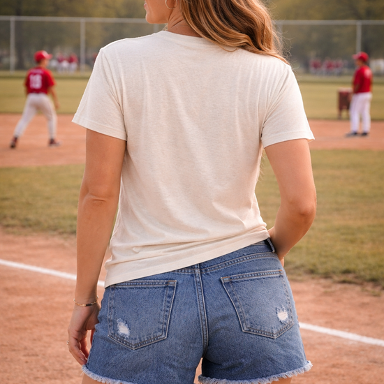 back view of woman wearing neutral tis the season baseball graphic tee with distressed denim shorts at a baseball field