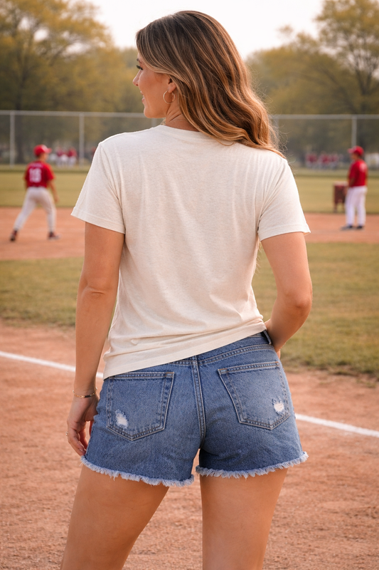 back view of woman wearing neutral tis the season baseball graphic tee with distressed denim shorts at a baseball field