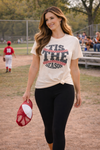 woman wearing tis the season baseball graphic tee with black leggings walking at a baseball field holding a cap