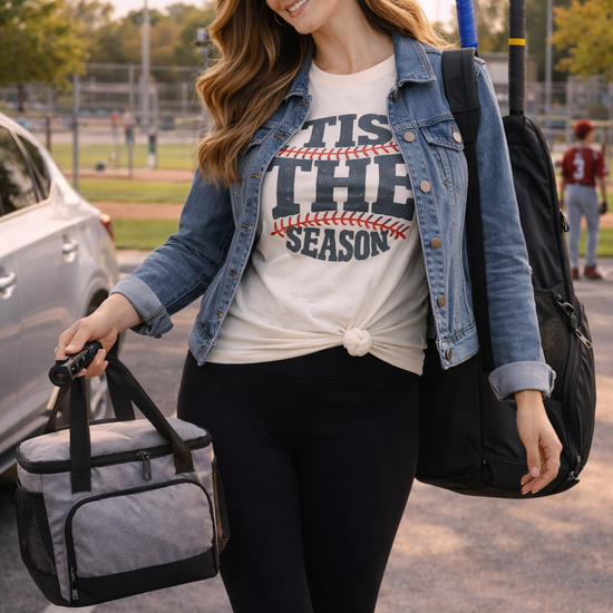 woman wearing tis the season baseball graphic tee with black leggings and denim jacket carrying cooler bag and bat bag walking near a baseball field