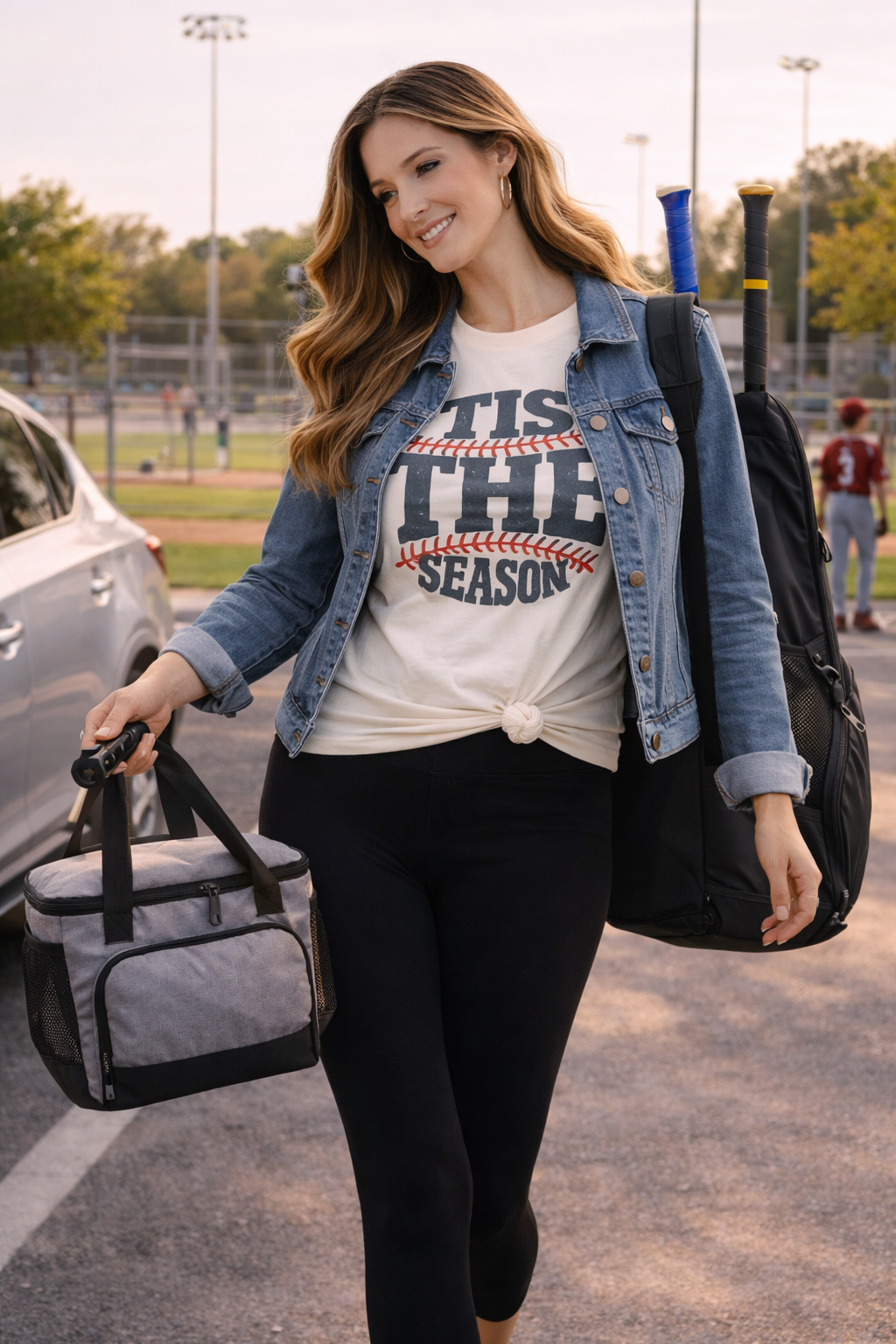 woman wearing tis the season baseball graphic tee with black leggings and denim jacket carrying cooler bag and bat bag walking near a baseball field