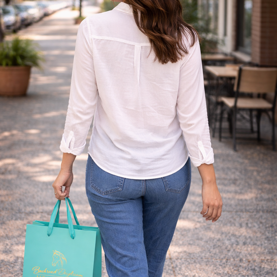 Back view of a woman walking outdoors wearing a white button-up blouse and holding a Backroad Daydream Mercantile shopping bag