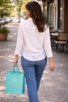 Back view of a woman walking outdoors wearing a white button-up blouse and holding a Backroad Daydream Mercantile shopping bag