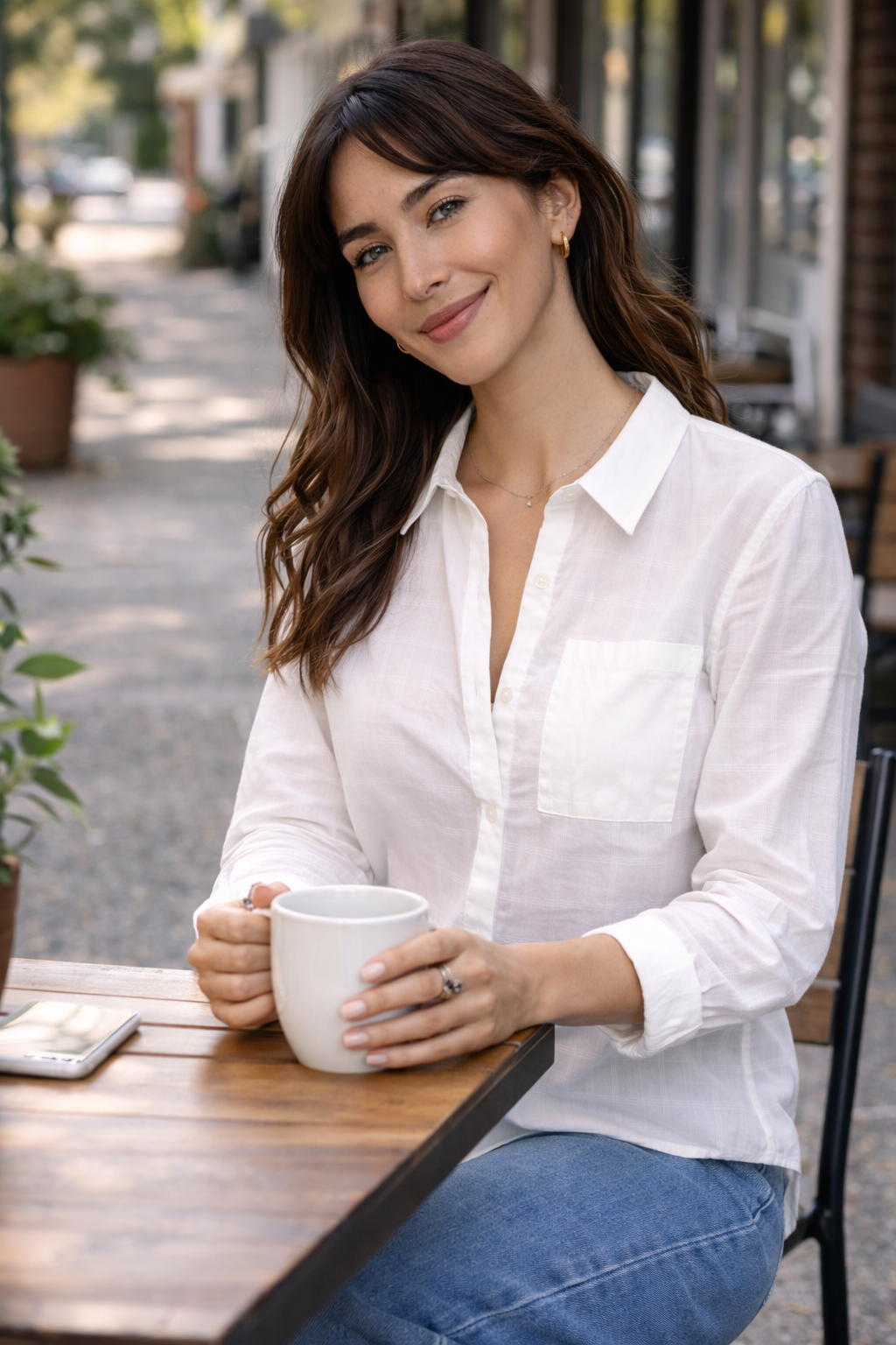 woman sitting at an outdoor café wearing a lightweight white button-up blouse styled with denim for a casual everyday outfit